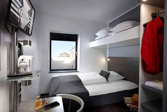 A compact cabin room with a bed under a window, an upper bunk and a red jacket hanging in an open closet.