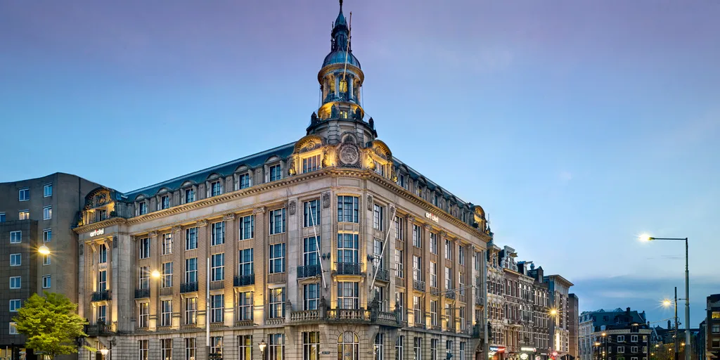 A corner hotel building with a small dome, lit at dusk on a city street.