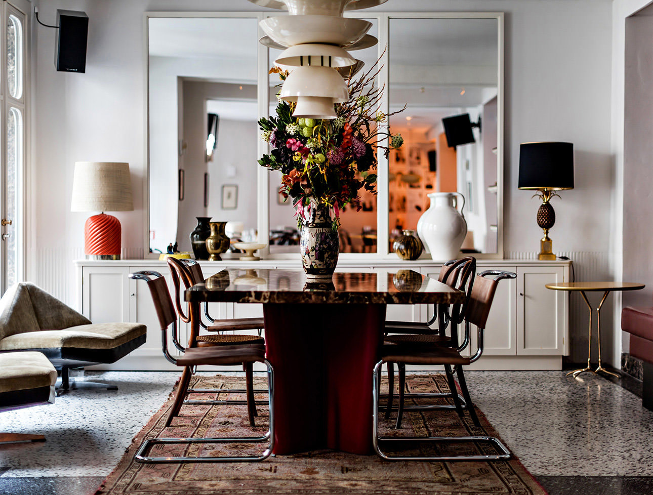 A dining room with a marble table, leather chairs, a tall flower arrangement and a long white sideboard.